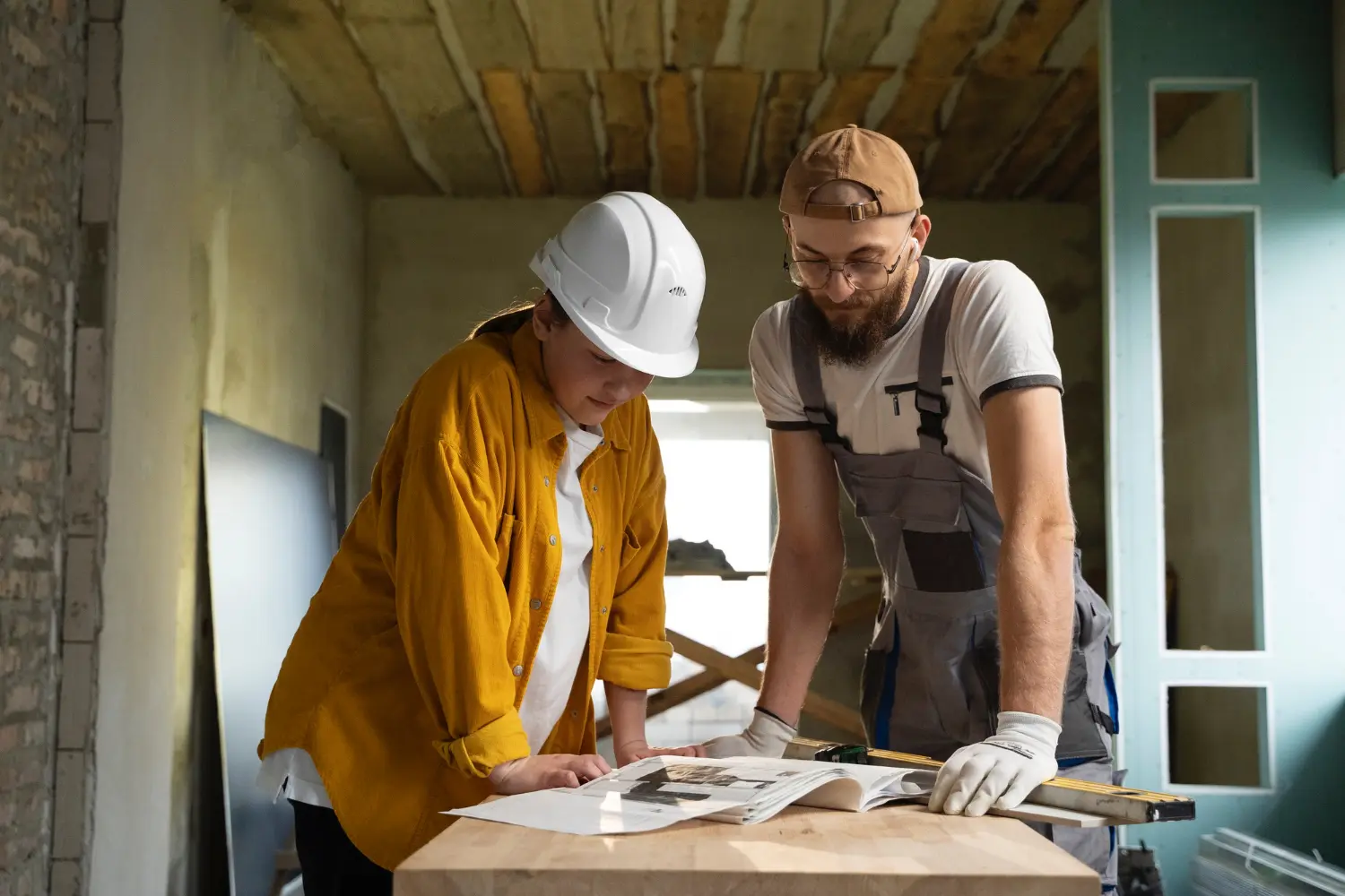 Homme et femme en tenue de chantier avec casque, consultant le plan d’une maison en rénovation intérieure avec plaques